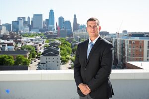 Man in a dark suit standing on a rooftop with the Kansas City skyline and a construction crane behind him
